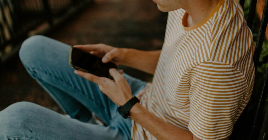 A person in a striped shirt and denim, sitting cross-legged, holds and looks at a smartphone.
