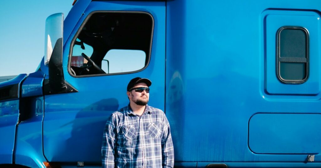 A man wearing a cap, sunglasses, and a plaid shirt stands in front of a large blue semi-truck cab, looking off to the side.