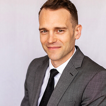Professional headshot of Cody J. Wagner, smiling, wearing a gray suit with a black tie, against a light gray background.