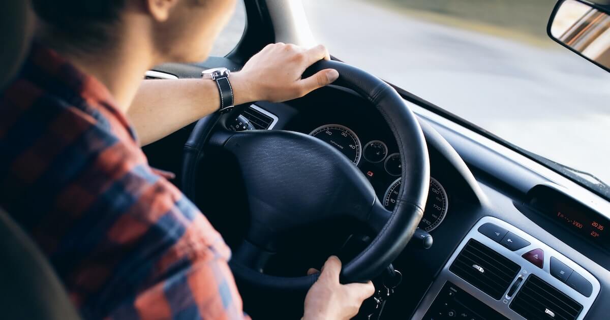 Over-the-shoulder view of a person driving a car, hands on the steering wheel, dashboard gauges visible.