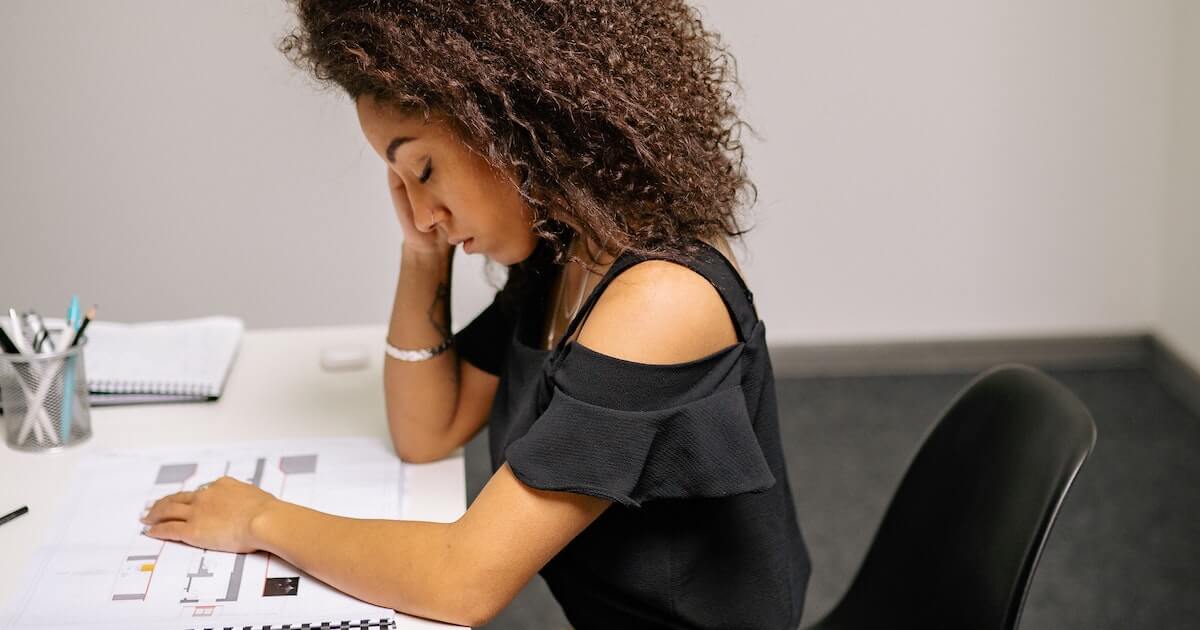 A woman with curly hair sits at a desk resting her head in her hand, looking down at architectural floor plans, appearing tired or stressed.