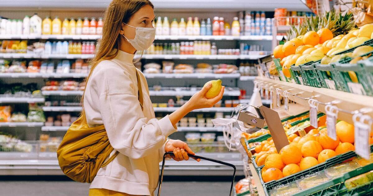 A woman wearing a face mask holds a lemon while shopping in a grocery store's produce section, with citrus fruit displayed on shelves nearby.