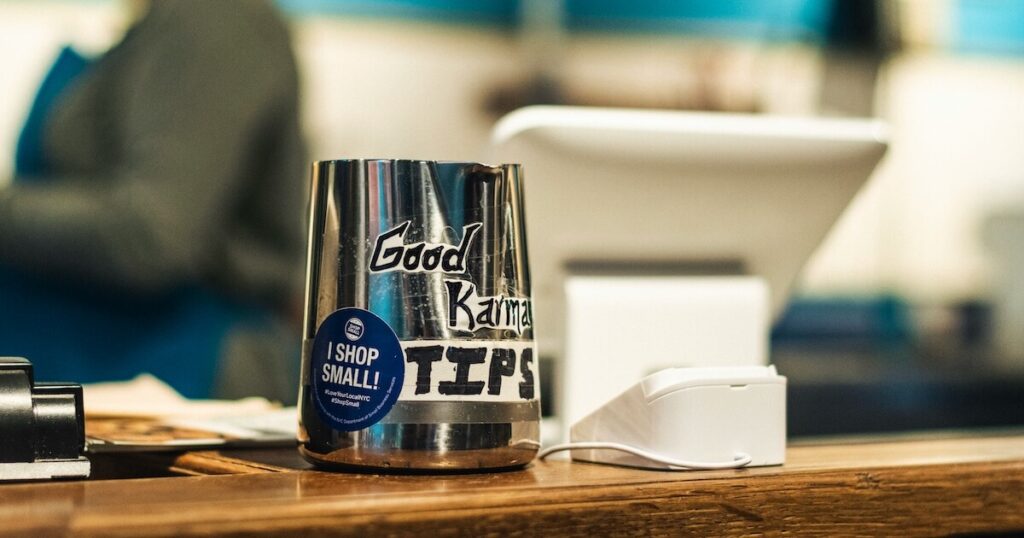 A metal tip jar labeled 'Good Karma Tips' with an 'I Shop Small' sticker, sitting on a wooden counter near a payment terminal.