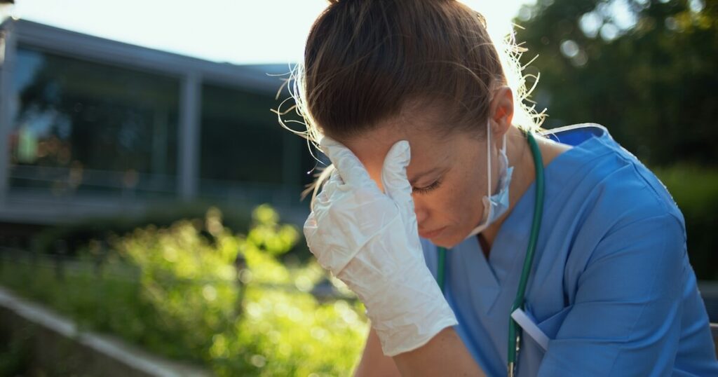 A healthcare worker in blue scrubs, wearing a stethoscope and a surgical mask pulled down, sits outdoors with her gloved hand pressed to her forehead, looking exhausted.