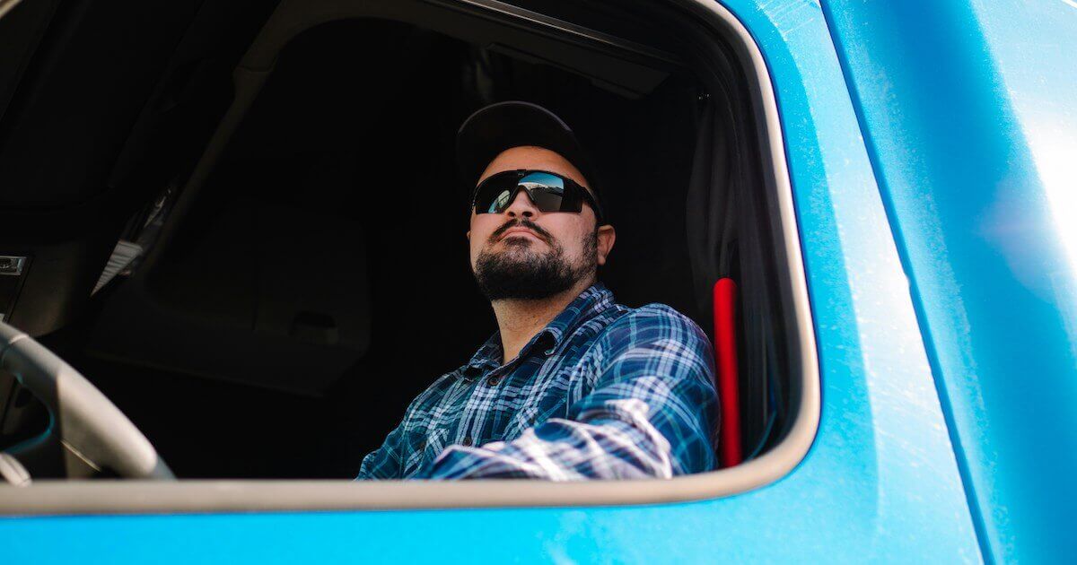 A man wearing sunglasses and a cap sits behind the wheel of a blue truck, seen through the driver's side window.