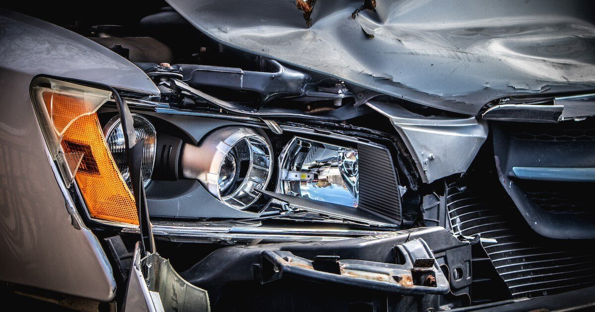 Close-up of a car's damaged front end after a collision, showing a crumpled hood and broken headlight assembly.