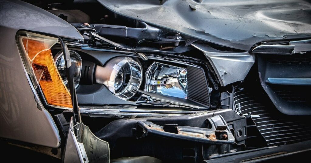 Close-up of a car's damaged front end after a collision, showing a crumpled hood and broken headlight assembly.