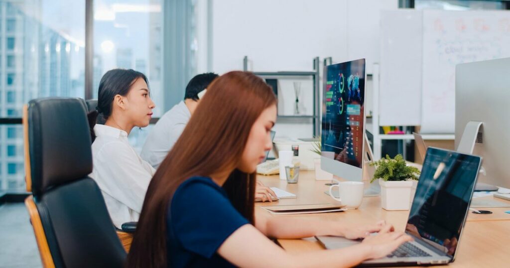 Three coworkers sit at a shared desk in a modern office, working on laptops and a desktop monitor displaying data charts, with a city skyline visible through the windows.