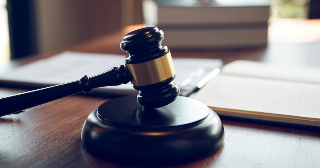 Close-up of a judge's gavel and sound block resting on a wooden desk, with an open book and stacked papers in the background.