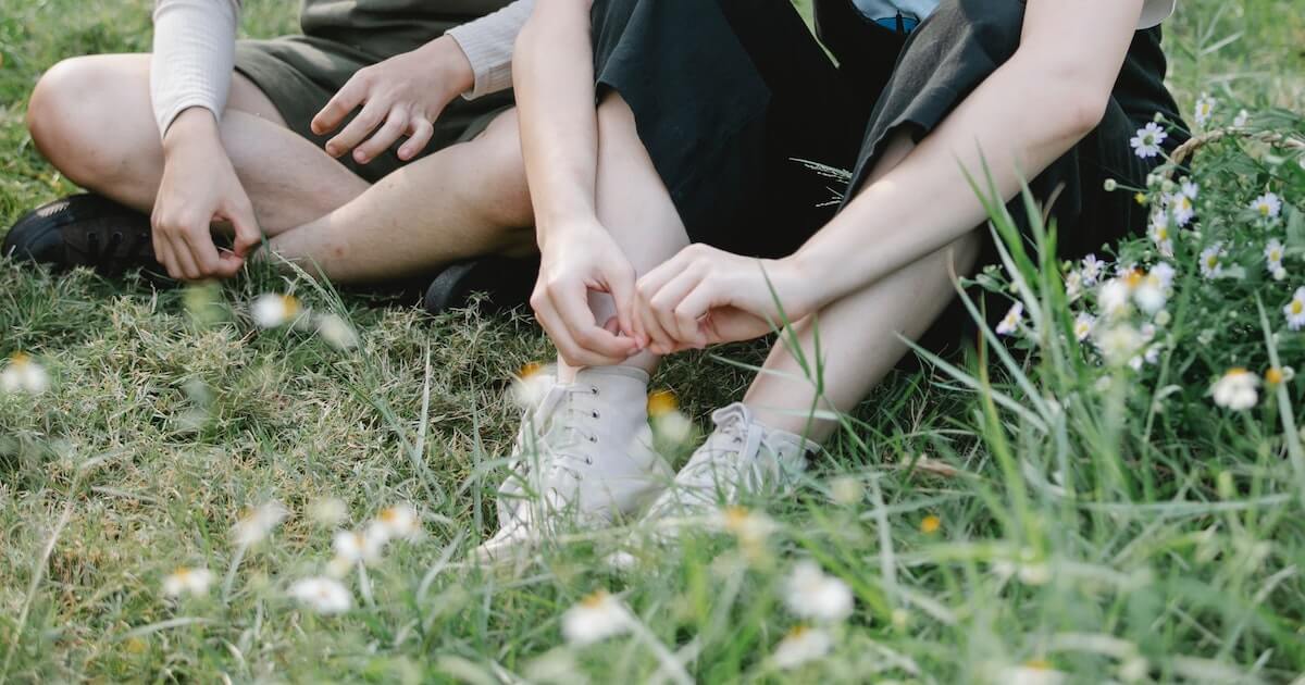 Two people sit cross-legged in a grassy field dotted with small wildflowers, their legs and hands visible in close-up as one picks at flowers near their sneakers.
