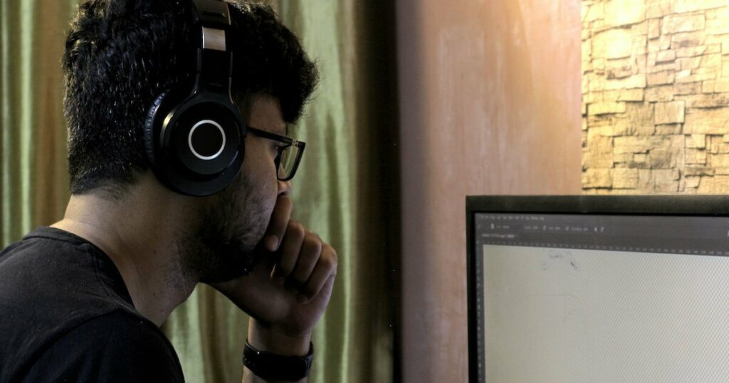 A man wearing headphones and glasses studies a large blank computer monitor, resting his chin on his hand in thought.