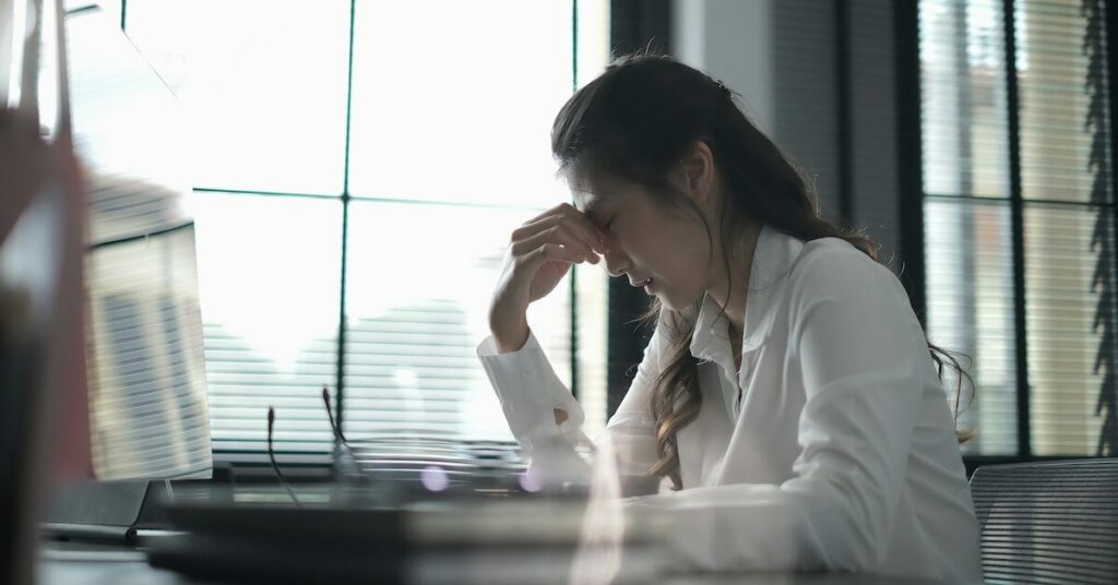 A woman in a white blouse sits at a desk pinching the bridge of her nose, looking stressed, with a bright window and blinds behind her.