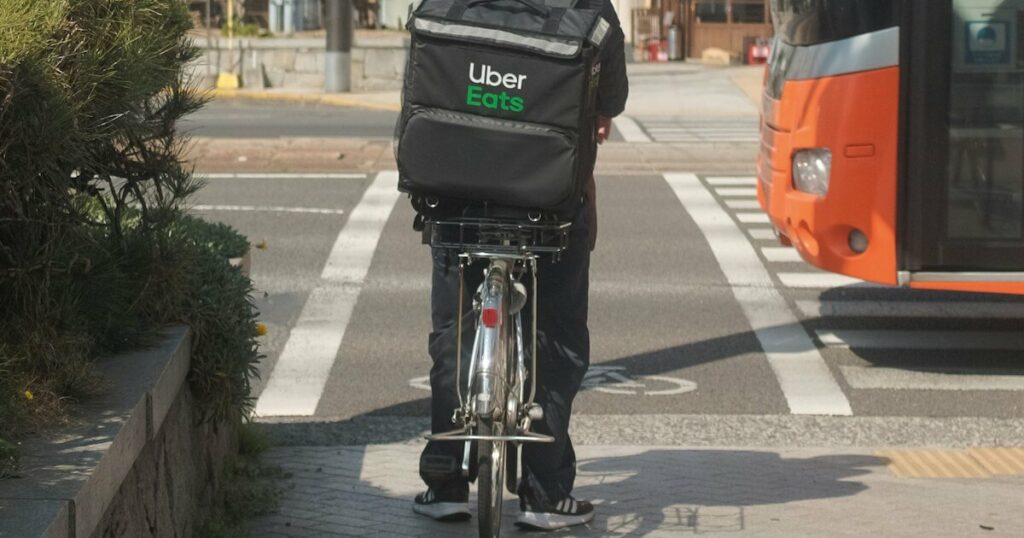 Rear view of a food delivery cyclist with an Uber Eats bag on his back, waiting to cross a street next to an orange bus.