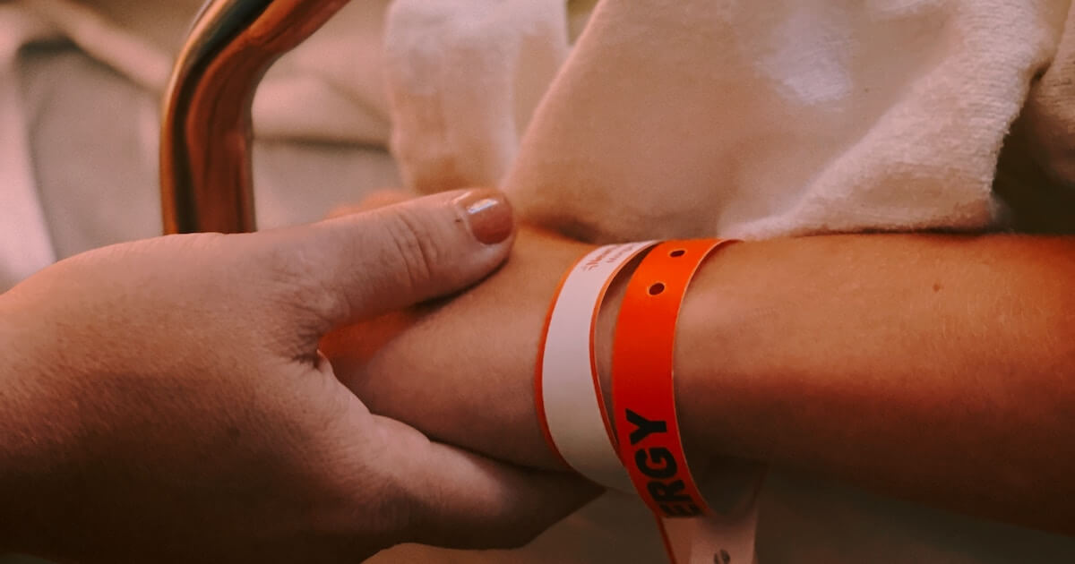 Close-up of a hand holding a patient's wrist, which has an orange allergy alert wristband and a white hospital ID band.