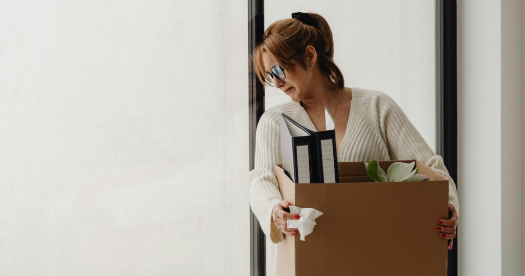 A woman wearing glasses and a cream sweater holds a cardboard box containing binders and a plant, looking down with a tissue in hand, as if leaving an office after being let go.