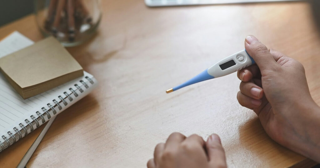 Close-up of a person's hands at a desk holding a digital thermometer, with a notepad and jar of pencils nearby.