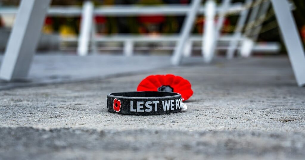 A black wristband engraved with a poppy symbol and the words 'Lest We For...' lying on pavement next to a red poppy pin.