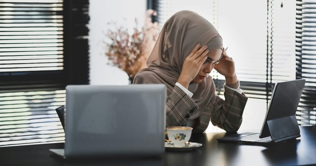 A woman wearing a hijab and blazer sits at a desk with her hands on her temples, looking stressed while working between a laptop and tablet, with a teacup nearby.