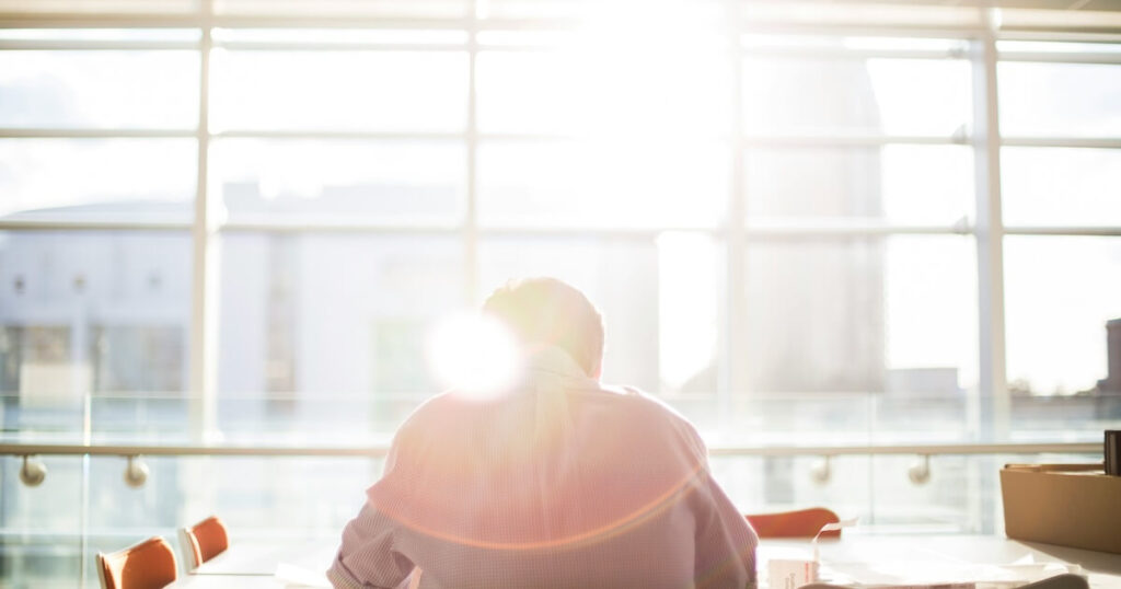 Silhouette of a man from behind, seated at a table in a brightly sunlit room with large windows overlooking a city.
