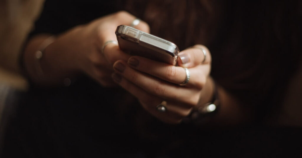 Close-up of a person's hands, adorned with several rings, holding and using a smartphone in low light.