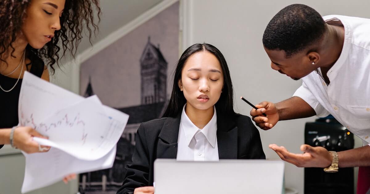 A woman sits at a laptop with her eyes closed and an overwhelmed expression, while two coworkers lean in on either side of her, one holding papers with charts and the other gesturing and pointing a pencil near her.