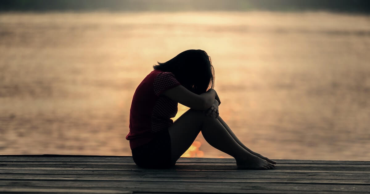 Silhouette of a person sitting on a wooden dock at sunset, knees pulled up and head resting on their arms, facing the water.
