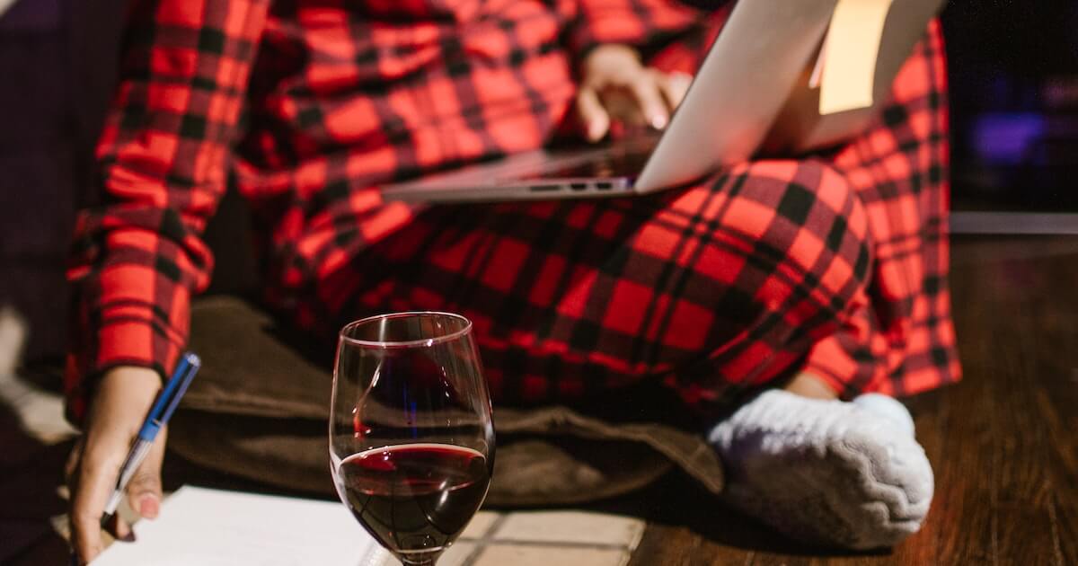 A person in red plaid pajamas sits cross-legged working on a laptop late at night, with a glass of red wine and notepad on the table in front of them.
