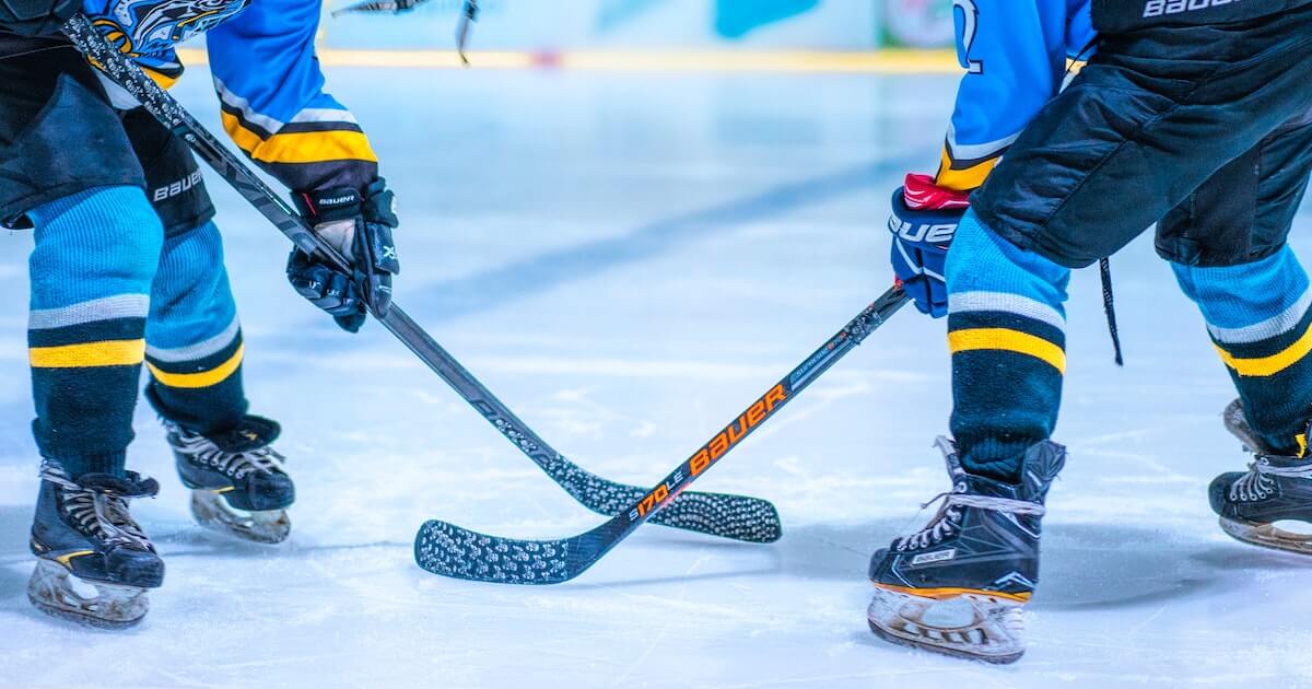 Close-up of two hockey players' sticks crossed at a faceoff on the ice, with their skates and lower legs visible.