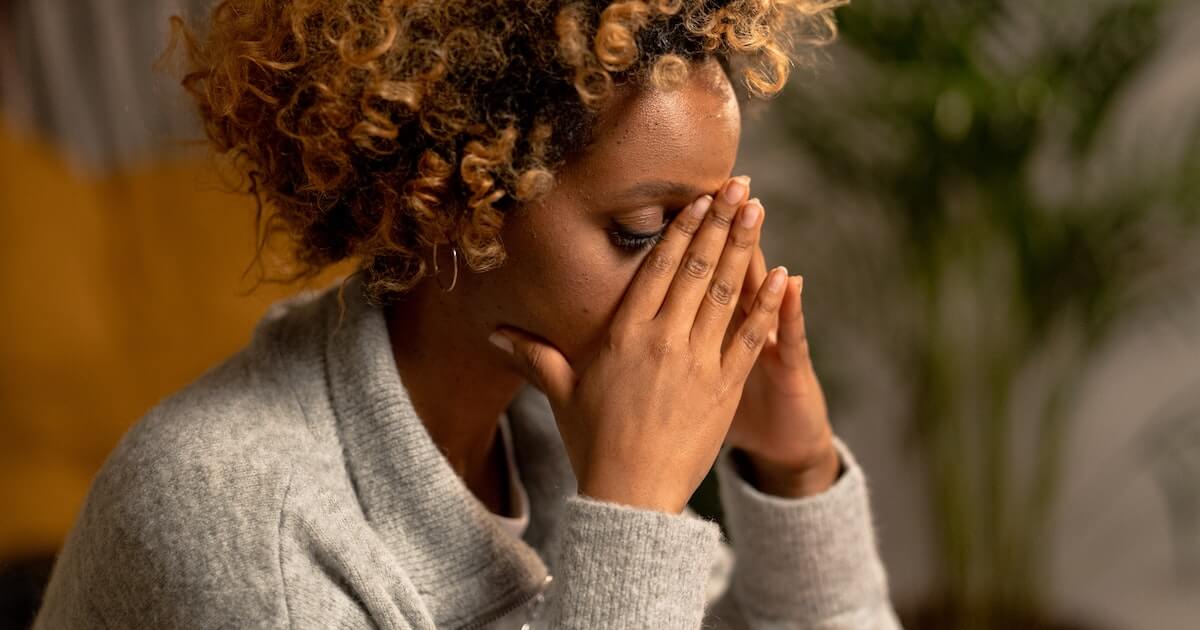"A woman with curly hair covers her face with both hands, appearing distressed, seated indoors with a plant in the background.