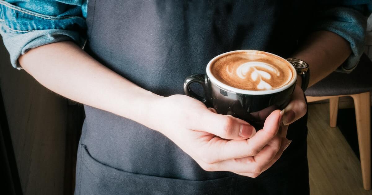A barista wearing a denim shirt and dark apron holds out a black cup of coffee with latte art, offering it to the camera.
