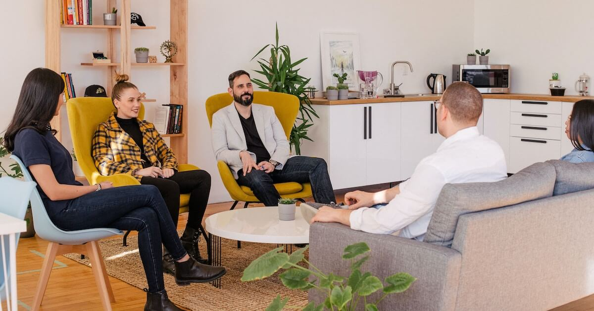 A group of five colleagues sit together on chairs and a couch in a casual, plant-filled lounge area, engaged in conversation.