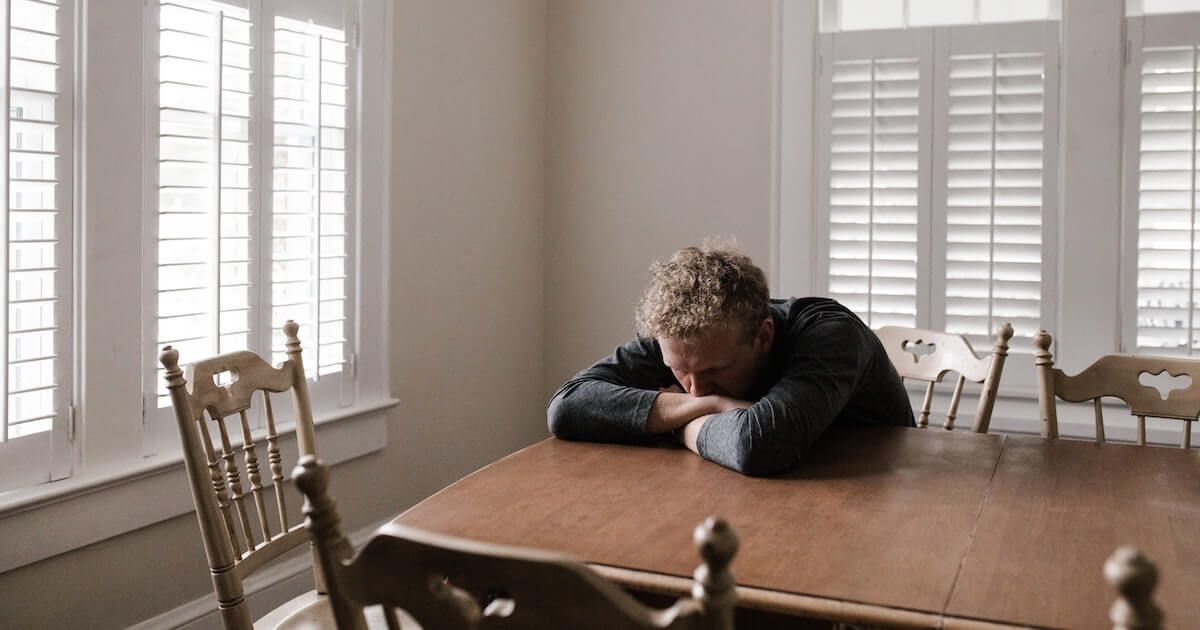 A man rests his head on his folded arms at a wooden dining table, looking exhausted or upset, with bright shuttered windows in the background.