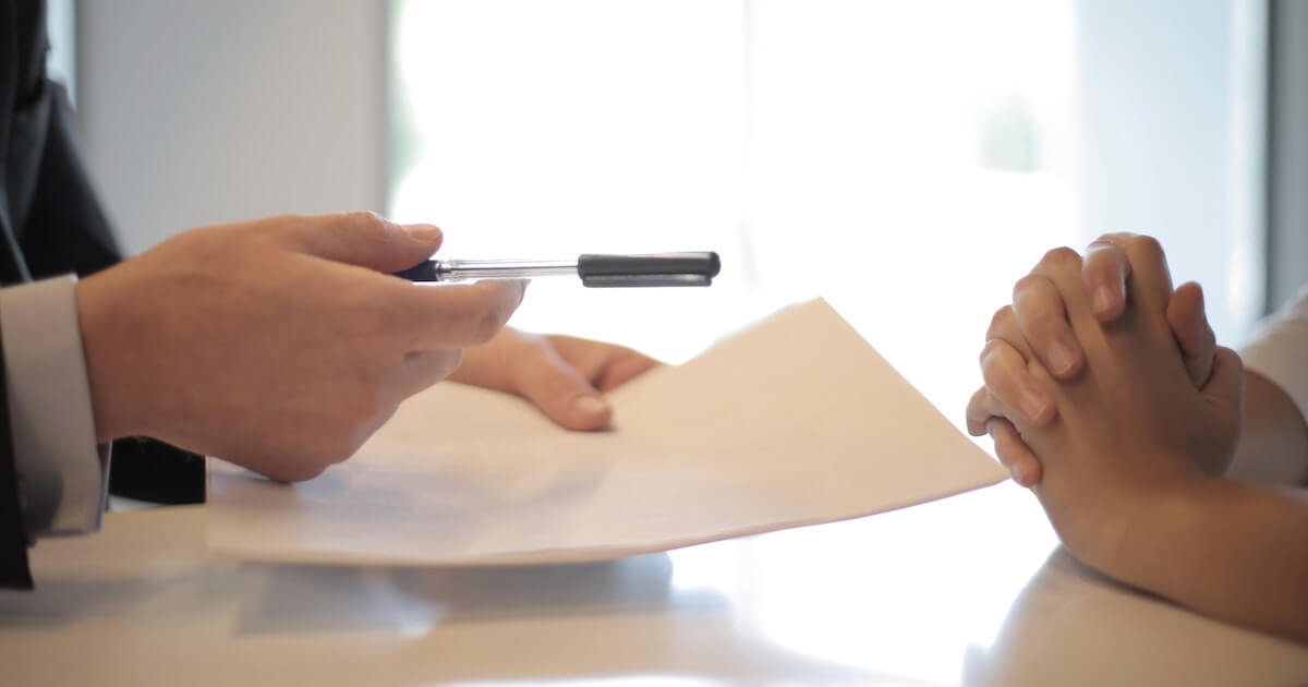One person holds out a pen and document across a table toward another person, whose hands are clasped, suggesting a contract signing.
