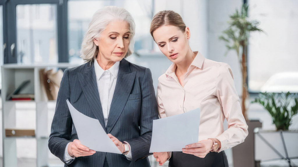 Two businesswomen stand reviewing documents together in an office, one with gray hair in a pinstripe suit and the other in a light blouse.