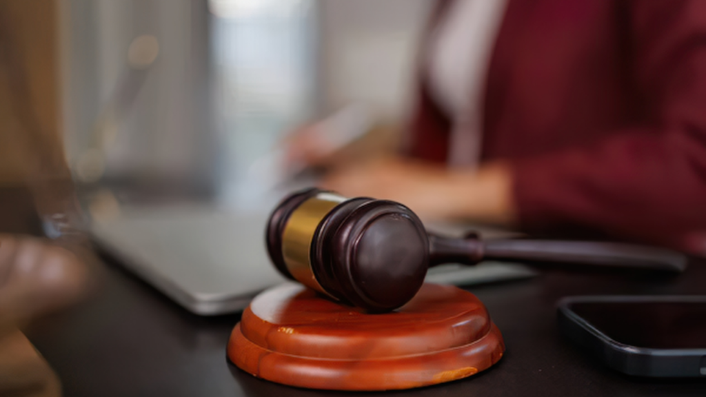 Close-up of a wooden gavel and sound block on a desk, with a person working on a laptop in the blurred background.