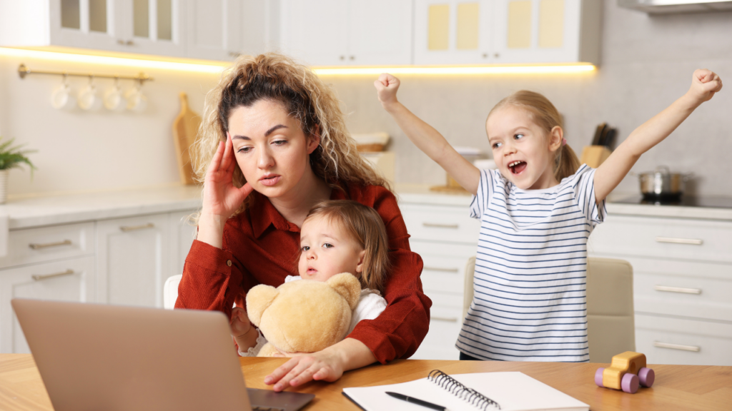 A stressed woman sits at a laptop holding a toddler and a teddy bear, while an older child excitedly raises her arms in a kitchen.