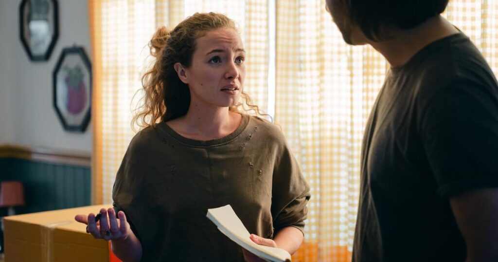 A woman with curly hair speaks to a man with a concerned expression, standing indoors near a moving box and sheer curtains, holding a folded piece of paper.