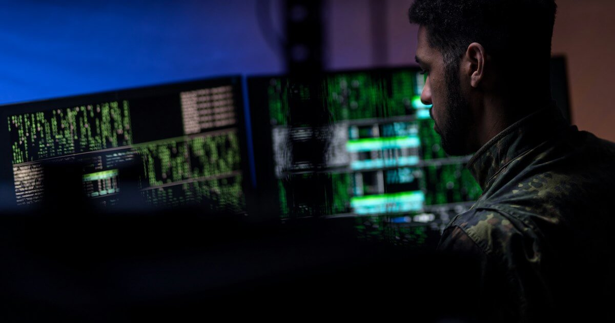 A person in military camouflage attire is silhouetted against multiple computer monitors displaying green and white data readouts in a dark room.