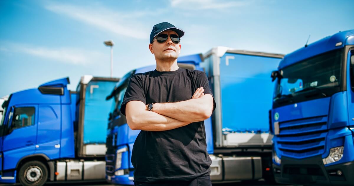 A man in sunglasses and a cap stands with his arms crossed in front of a fleet of blue semi-trucks.