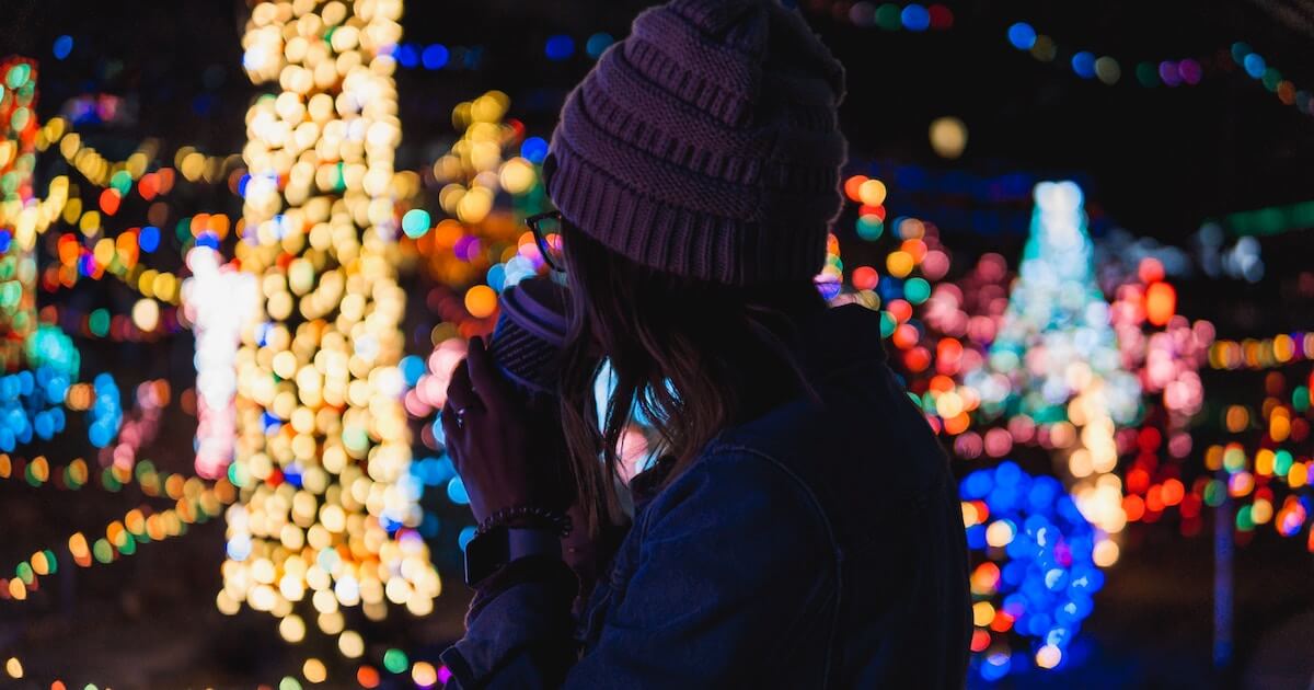 Silhouette of a person wearing a beanie, seen from behind holding a drink, surrounded by blurred colorful string lights at night.