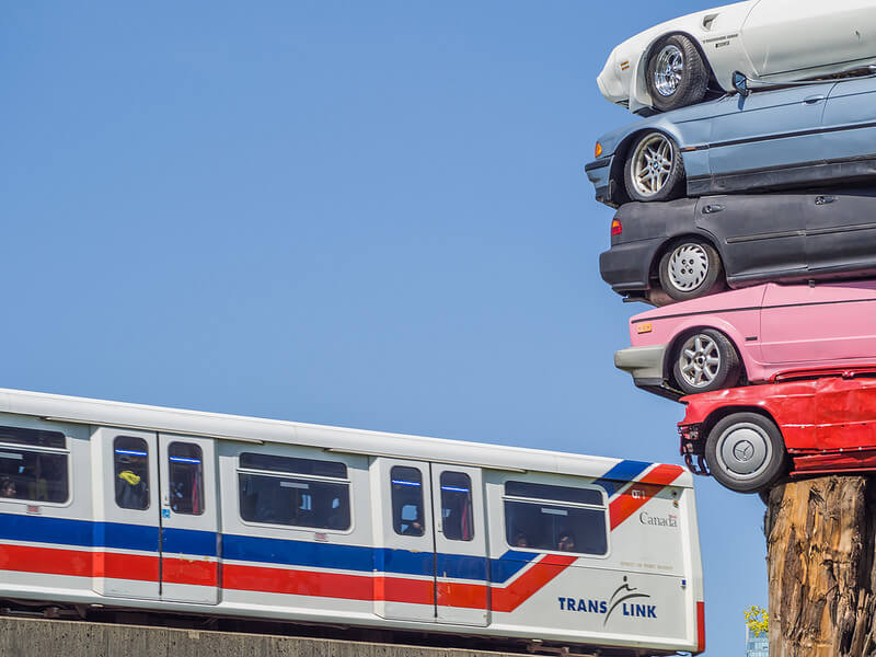 A train going past an art display of five cars stacked on top of each other