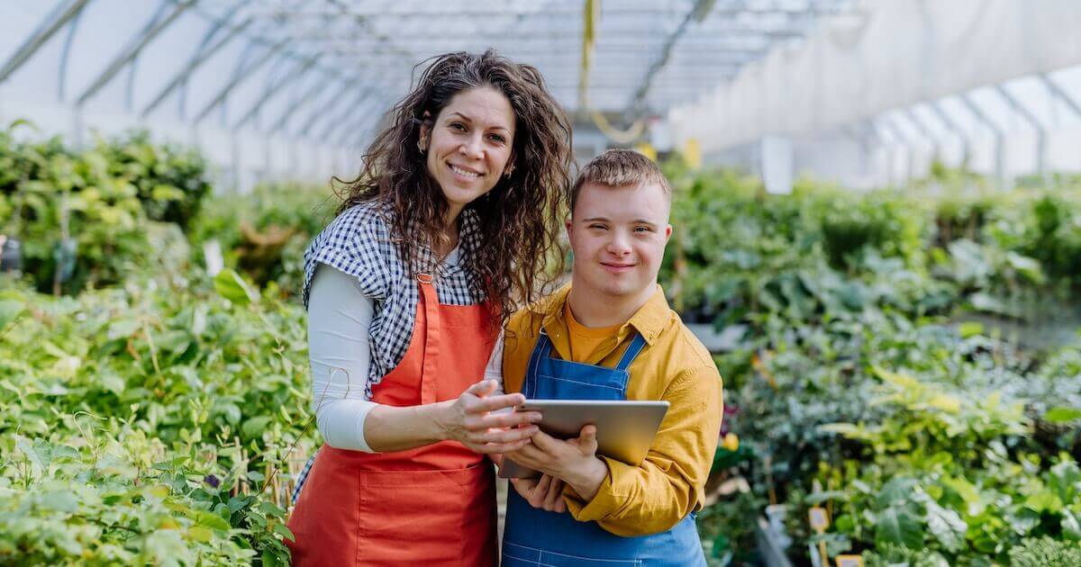 Two side-by-side colleagues view an iPad screen together while standing inside a large greenhouse