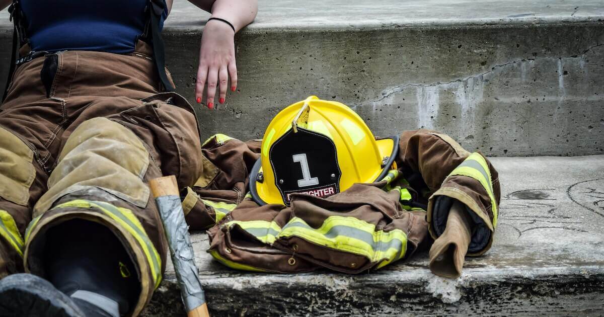 A female firefighter rests against a concrete ledge with her turnout coat and bright yellow helmet resting beside her