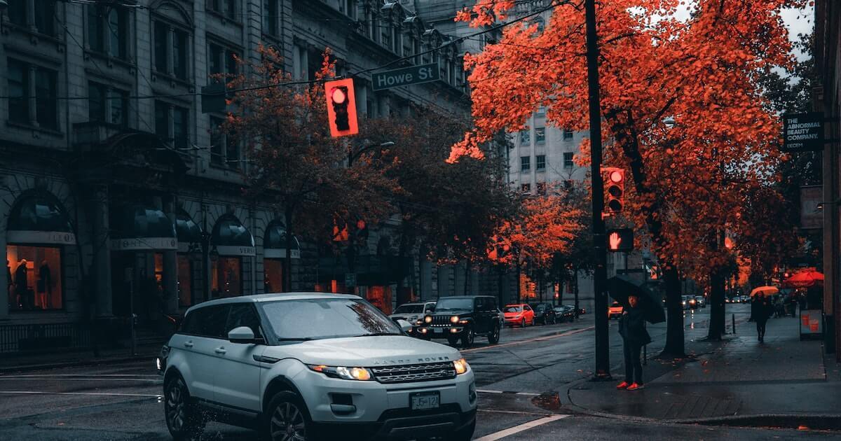 A white Range Rover turns the corner of a downtown Vancouver street lined with crimson maple trees