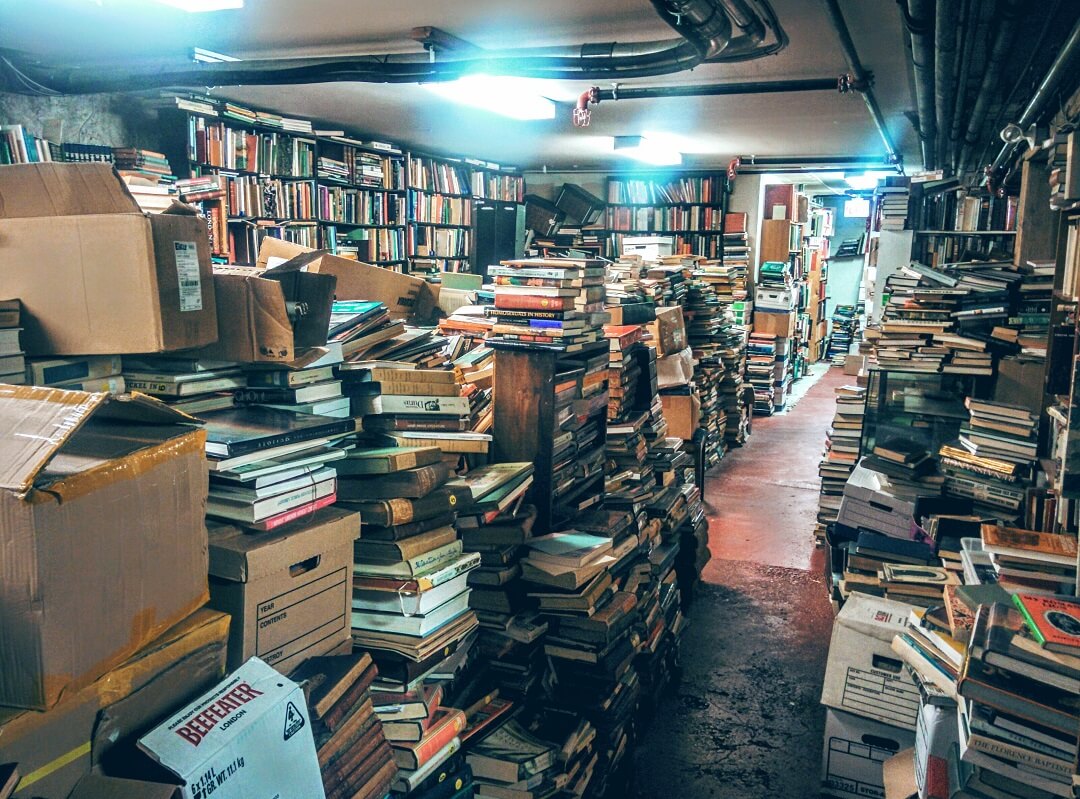 A room with books lining the shelves and piles in the middle of the room