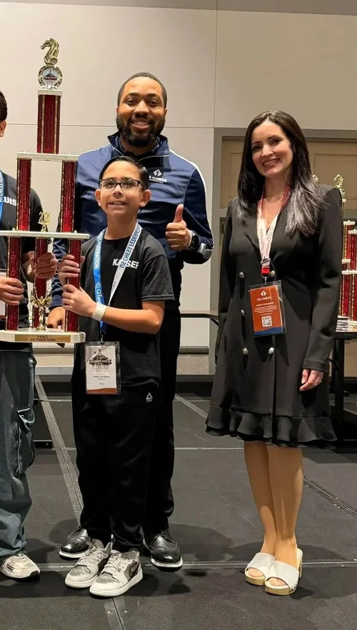 Proud mother standing on stage supporting her young son holding a large chess trophy.  Shared by RGV Chess and Learning Center.