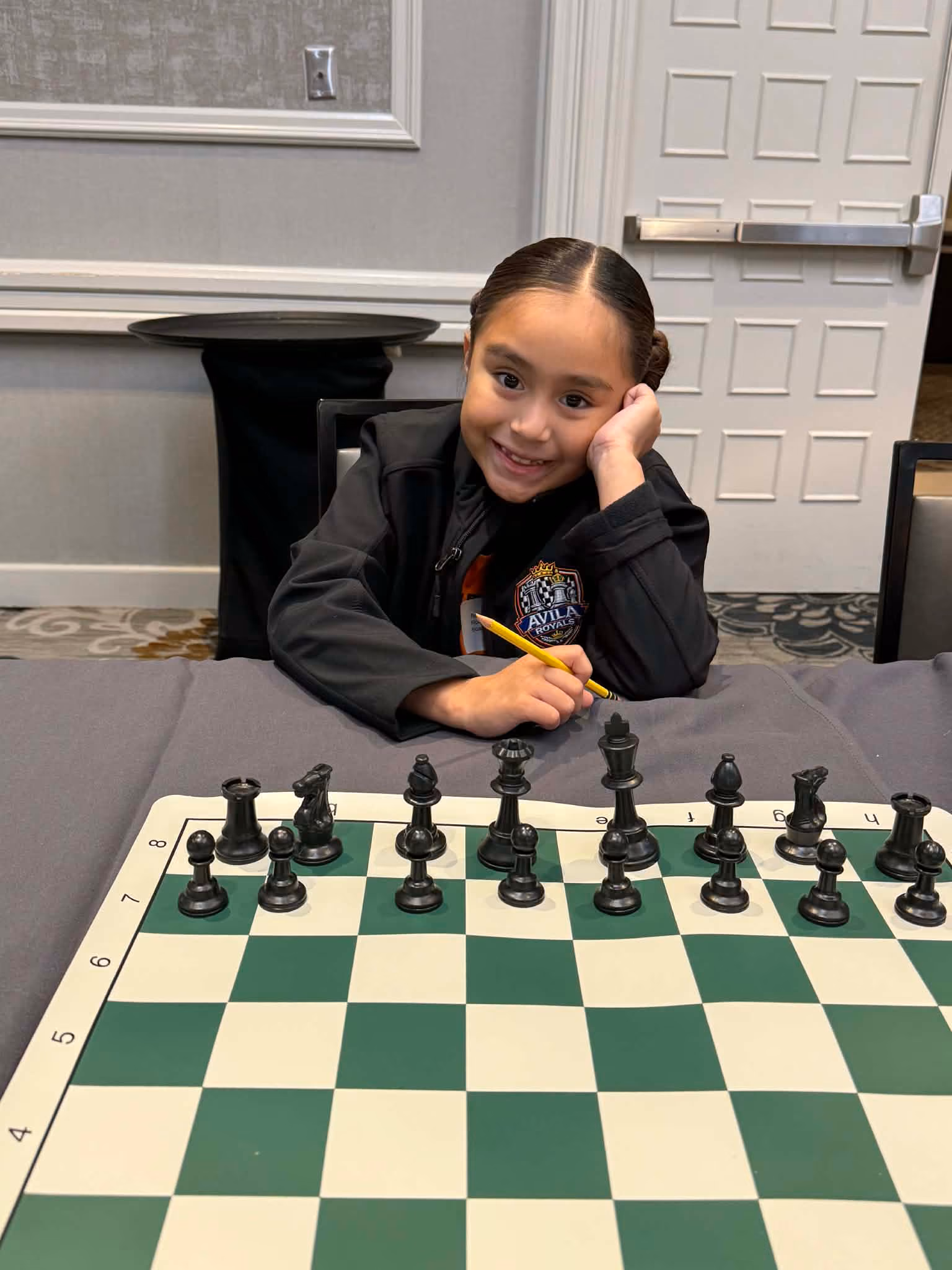 Smiling elementary school student sitting at a tournament chess board preparing to play and take notation. Shared by RGV Chess and Learning Center.