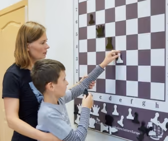 An instructor supporting a student with autism as he places a piece on a large demonstration board during an adaptive lesson. Shared by RGV Chess and Learning Center.