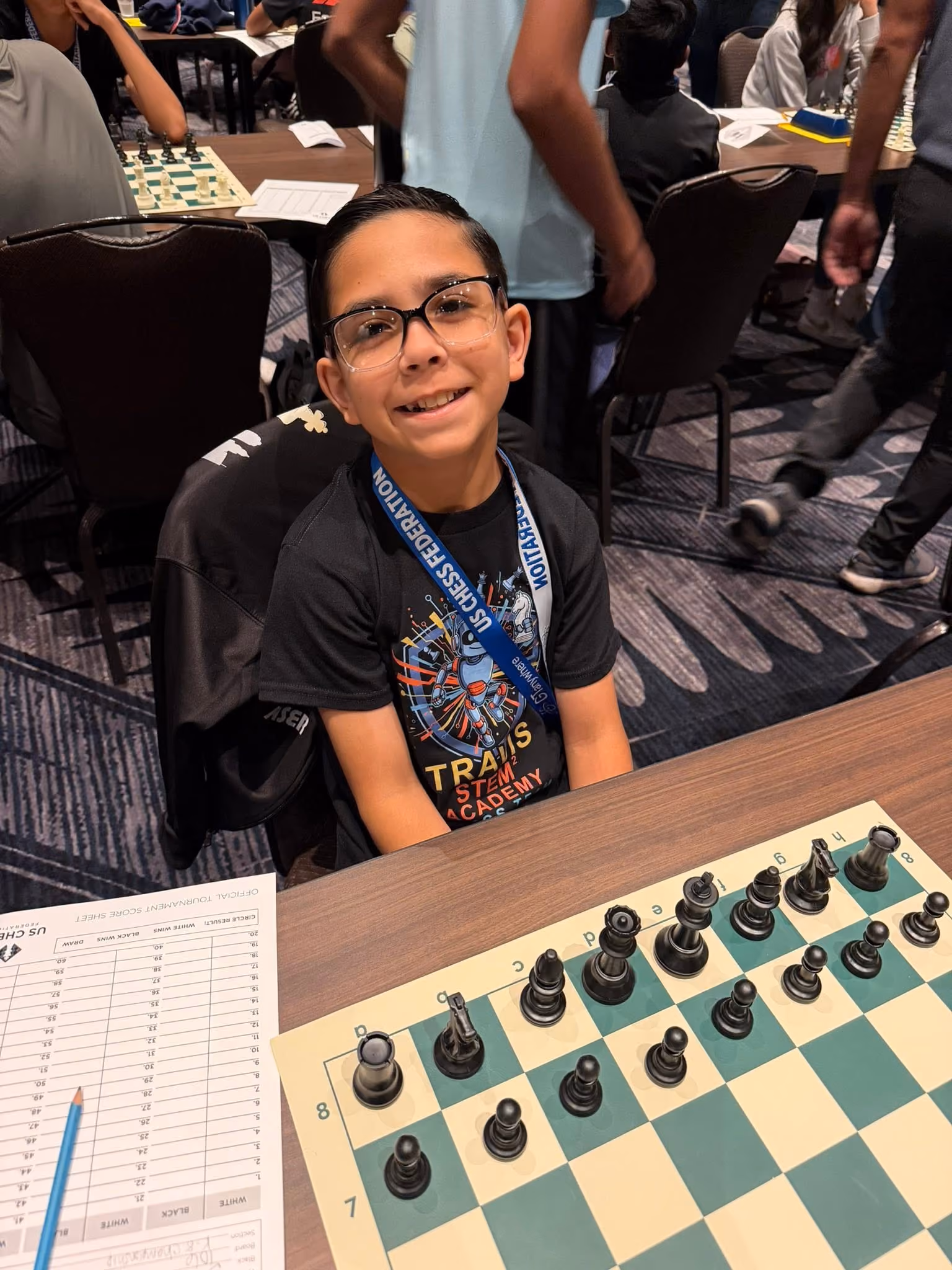 Elementary school student wearing a STEM shirt and US Chess lanyard sitting behind black pieces during a tournament shared by RGV Chess and Learning Center.