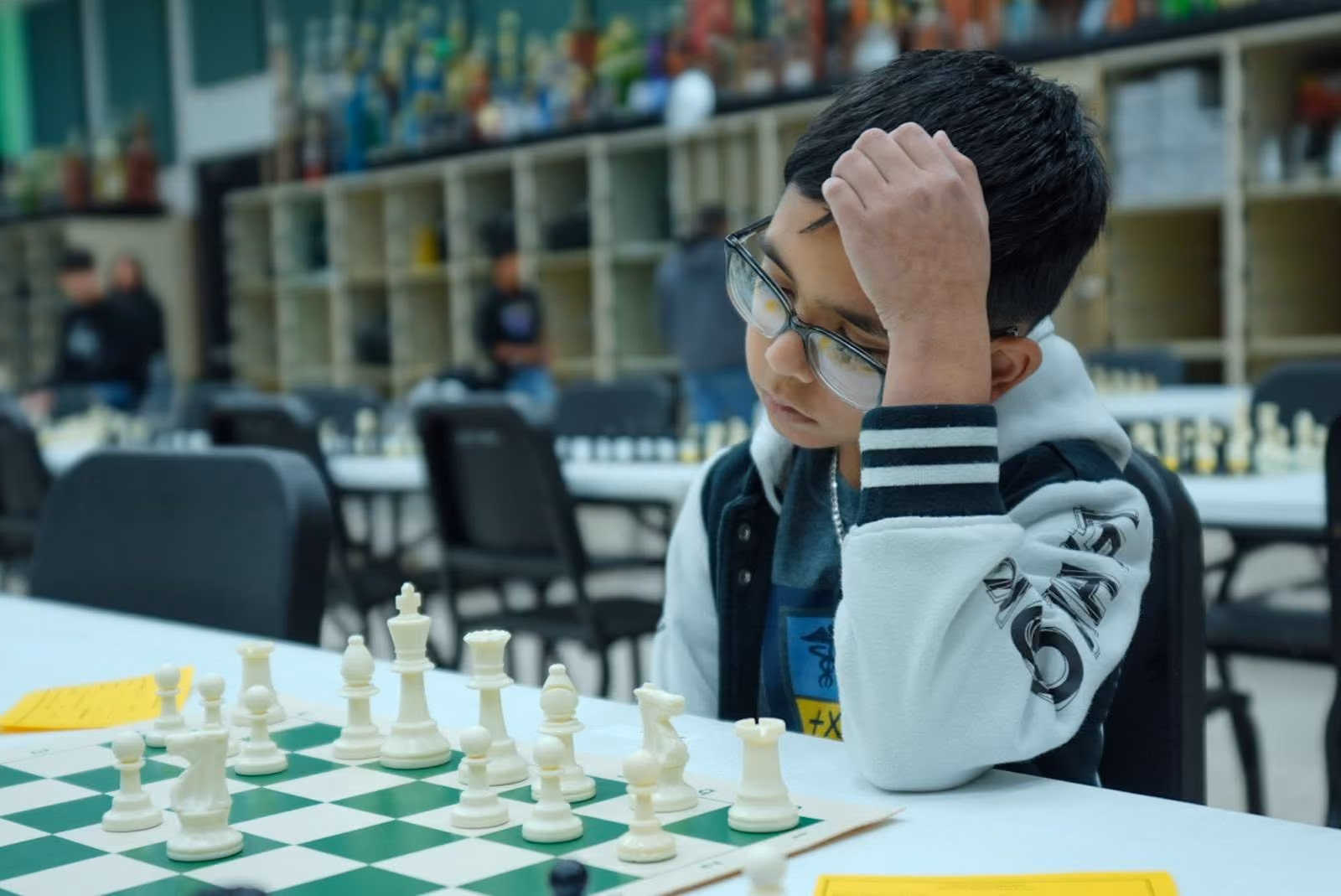 A close-up photograph of a young boy with his hand on his head, intense focus in his eyes, examining a chess game in progress at a tournament.